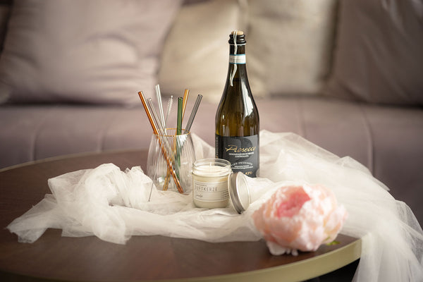 Bunte Glastrinkhalme im Glas mit einer Reinigungsbürste auf einem runden Holztisch, dekoriert mit einem weißen, drappierten Schleier, einer Duftkerze, Prosecco Flasche und mit einem Blütenkopf Pfingstrose in Rosa.