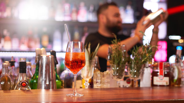 Theke mit einem Longdrink-Getränk in einem Weinglas mit einem klaren Glashalm. Im unscharfen Hintergrund ein Barman, der ein Cocktail mixt.