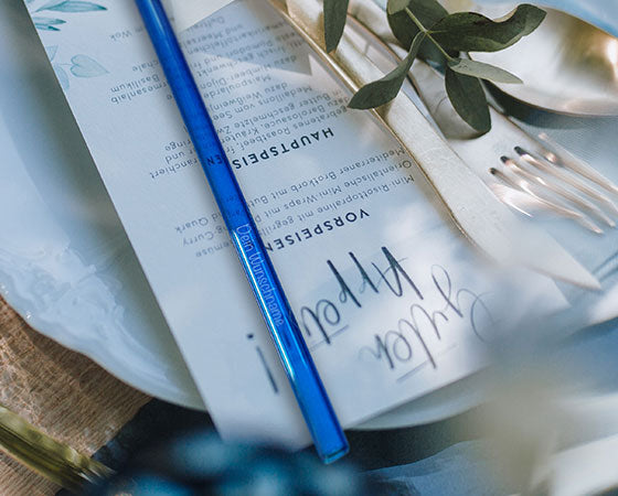 Ein festliches Gedeck mit Menükarte und Glastrinkhalm in Blau mit gravierten Schriftzug "Dein Wunschname"